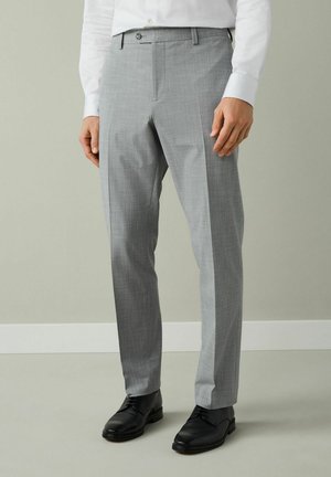 Vyras dėvi šviesiai pilkas kostiumines kelnes, baltą marškinius ir juodus odinius batelius, stovi priešais paprastą pilką foną.