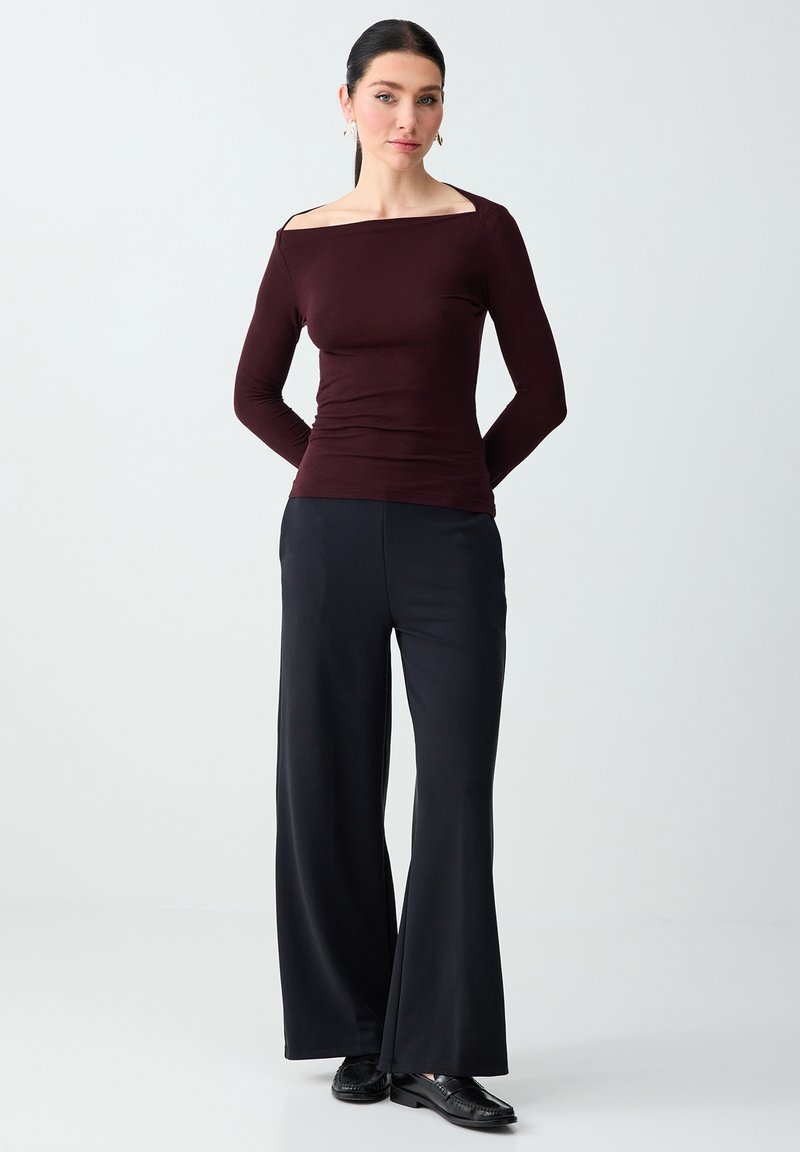 Femme debout, les mains derrière le dos, portant un haut bordeaux foncé décolleté aux épaules, un pantalon large noir et des mocassins noirs.