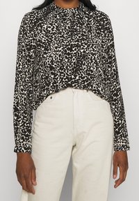 Blusa a maniche lunghe in stampa leopardata nera e beige, con un colletto alto e dettagli arricciati sulle spalle. Abbinata a pantaloni bianchi.
