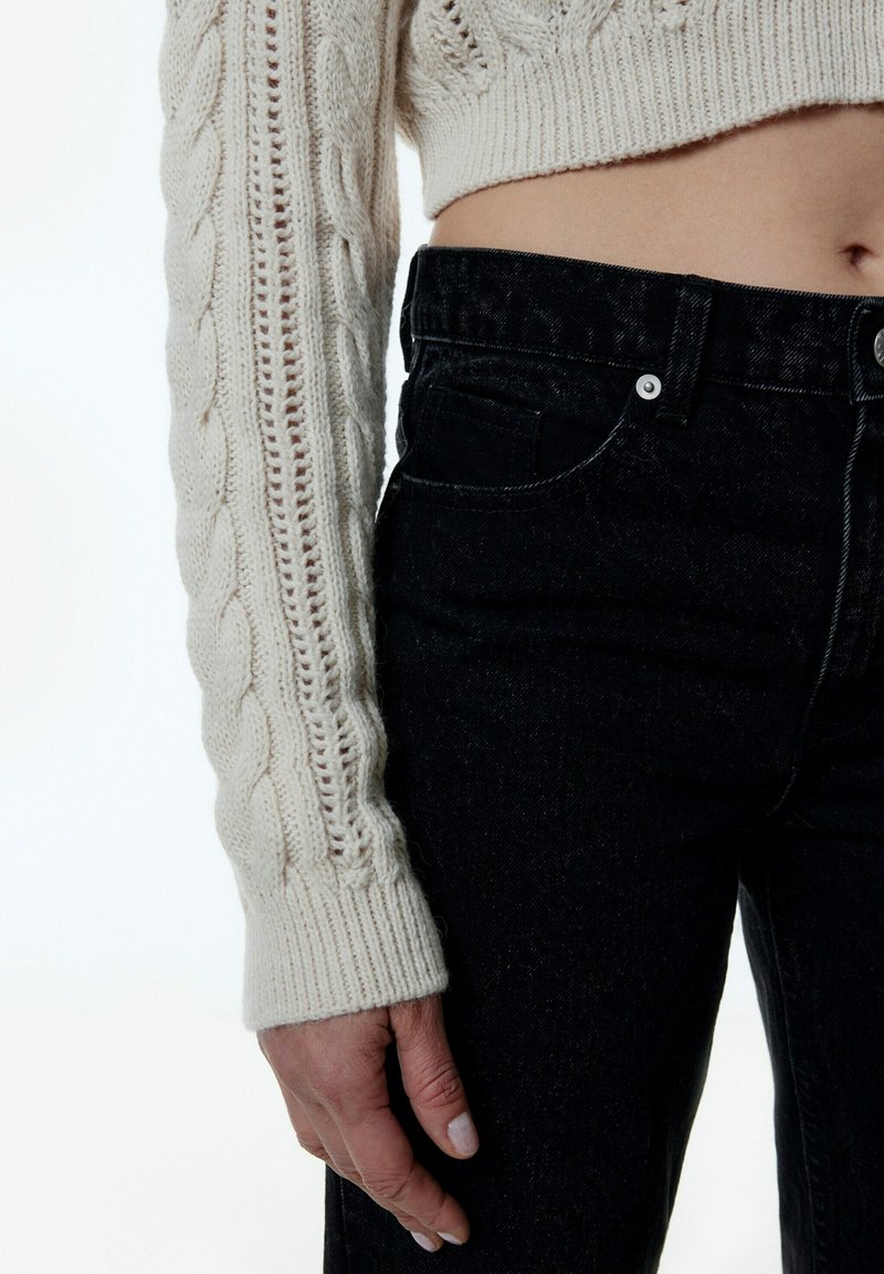 Pull en maille torsadée crème avec une coupe courte et des poignets côtelés, associé à un jean en denim noir avec une coupe classique à cinq poches.