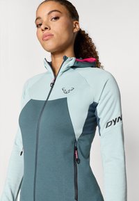 Kapuutsi sportlik jakk helesinine ja tumesinine, millel on lukuga sulgure, roosa aksent, tekstureeritud kangas ja logo varrukal.