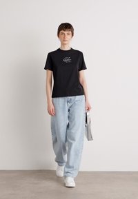 Zwarte katoenen t-shirt met een logo op de borst, gecombineerd met lichtblauwe wijde jeans en witte sneakers. Model houdt een zilveren tas vast.