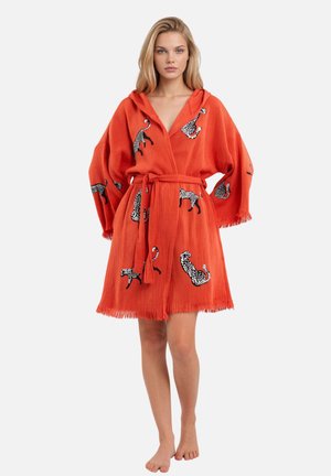 Vrouw draagt een losse oranje mantel met zwart-witte luipaardprints, gestrikt in de taille, staande op blote voeten tegen een effen achtergrond.