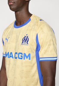 Κίτρινη αθλητική φανέλα με μπλε λεπτομέρειες, που διαθέτει υφή με σχέδιο τοποθεσιών ορόσημων, λογότυπο PUMA και τη λέξη "MACGM" με έντονα μπλε γράμματα.