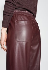 Pantaloni in finta pelle bordeaux con vita elastica e due tasche posteriori rettangolari che presentano una texture liscia e lucida.