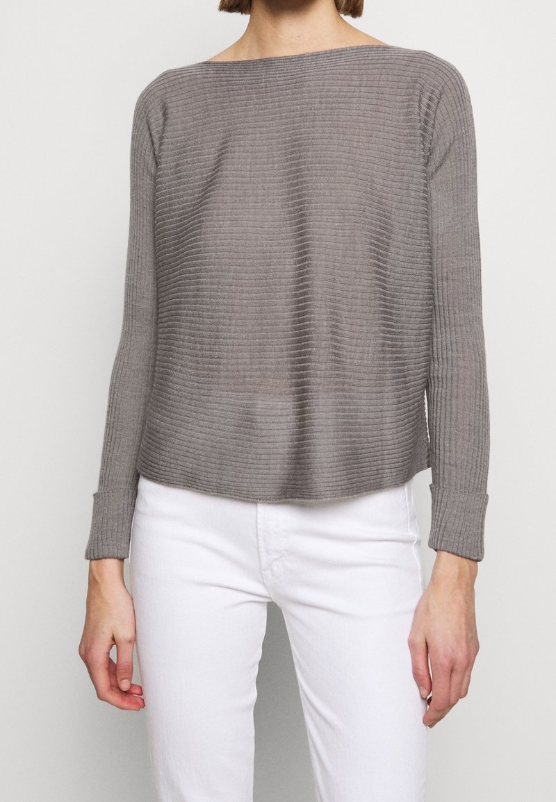 Pull gris côtelé avec un col bateau et des manches longues. Le tissu présente un aspect légèrement texturé et est associé à un pantalon blanc.