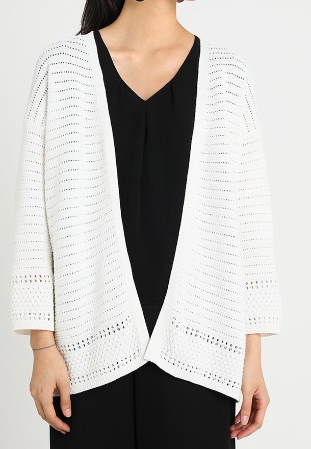 Vit öppen stickad cardigan med löst snitt, breda 3/4ärmar och ett detaljerat mönster vid ärmsluten och fållen, bärs över en svart topp.
