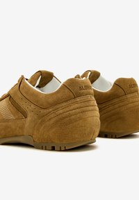 Le scarpe sportive in suede marrone presentano un pannello in rete, una punta arrotondata, dettagli testurizzati e una suola in gomma con un sottile dettaglio del marchio sul tallone.