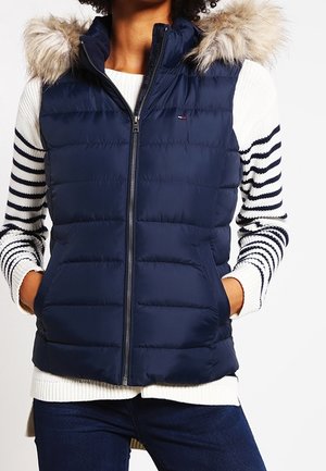 Femme portant une veste matelassée bleu marine avec une capuche bordée de fourrure par-dessus un pull blanc avec des rayures noires sur les manches.