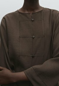 Haut en tissu marron avec col rond, fermetures par boutons et coupe ample avec manches larges. Design minimaliste avec surface texturée.