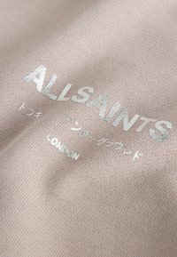 Hellrosa gewebter Stoff mit silberner, erhabener Prägung der Schriftzüge "ALLSAINTS" und "LONDON" sowie weiteren Zeichen darunter.