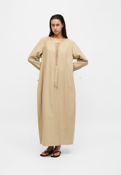 Femme portant une longue robe beige avec un détail à lacets sur l'avant, debout, les mains derrière le dos et portant des sandales noires sur un fond blanc uni.