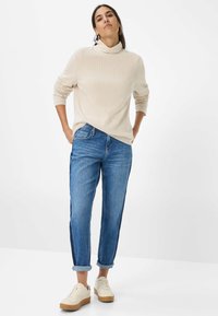 Lys beige strikket sweater med høj hals, parret med blå denimjeans med en sideliste. Kombineret med hvide sneakers.
