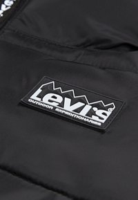 Crni pufasti kaput s ušivenim Levi's logom, s bijelim natpisom i dizajnom planine na vrhu. Glatka, mat tekstura.