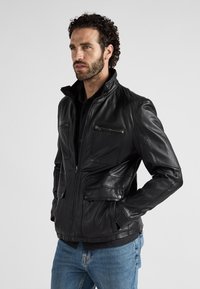 Schwarze Lederjacke mit hohem Kragen, Reißverschluss vorne, mehreren Taschen und strukturierten Akzenten. Verfügt über ein tailliertes Design und eine glatte Oberfläche.