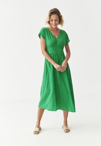 Grünes, knielanges Kleid mit V-Ausschnitt, kurzen Ärmeln und vertikaler Struktur. Ausgestellter Rock und definierte Taille, kombiniert mit neutralen Sandalen.