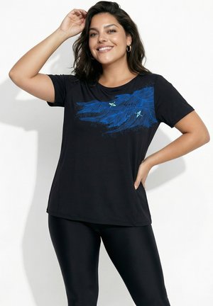Smilende kvinde med mørkt hår iført sort T-shirt med blåt fjer- og fugledesign, parret med sorte leggings, står mod hvid baggrund.