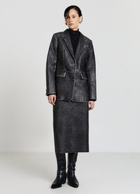 Blazer in ecopelle nera con cuciture a contrasto, abbinato a una gonna coordinata. La modella indossa stivali neri lucidi fino al ginocchio.