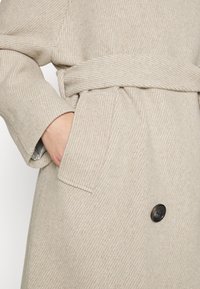 Beige geribbelde jas met een ceintuur in de taille, zijzakken en een enkele zwarte knoop. De stof heeft een zachte, gestructureerde uitstraling.