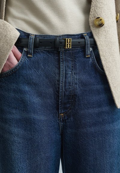 Σκούρο μπλε τζιν με ζώνη με πόρπη σε χρυσό χρώμα με λογότυπο, σε κανονική εφαρμογή και διακριτή υφή, συνδυασμένα με ένα μπεζ σακάκι.