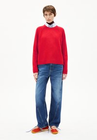 Rode gebreide trui, relaxte fit, ronde hals; gecombineerd met wijdvallende blauwe jeans; rood-oranje sneakers met witte veters en blauwe accenten.