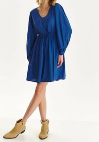 Sininen polvipituinen mekko, jossa on pitkät puff-hihat, vyötäröä korostava leikkaus ja V-pääntie; yhdistettynä vaaleanruskeisiin nilkkureihin. Sujuva kangasrakente.