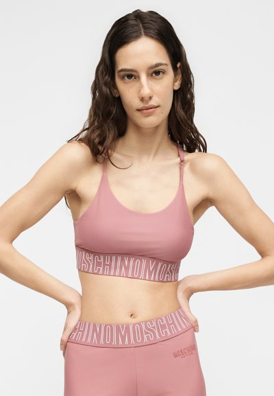 Νεαρή γυναίκα με μακριά κυματιστά μαλλιά που φοράει ροζ αθλητικό σουτιέν και κολάν Moschino με λαστιχένιες μπάντες με λογότυπο, τα χέρια ακουμπούν στους γοφούς.