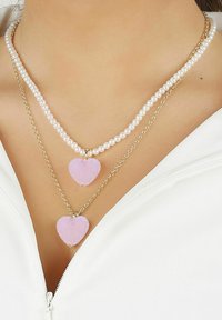 Dubbel-lager halsband: en fast rad av vita pärlor och en nedre kedja av guld med två rosa hjärtan, som har en glittrig yta.