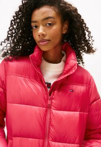 Red puffer jacket with a high collar, featuring a zippered front and a small logo on the chest. Smooth, glossy texture and quilted design.