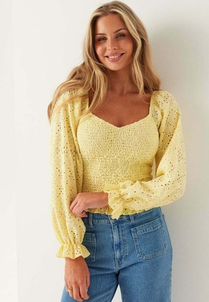 Ung kvinna med långt blont hår bär en gul blus med puffärm av hålmönster och blå högmidjade jeans, ler mot en enfärgad bakgrund.