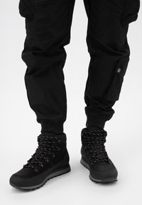 Černé turistické boty s texturovaným svrškem, zesílenou podrážkou a vzorovanými tkaničkami. V kombinaci s černými cargo kalhotami s bočními kapsami.