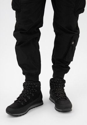 Zwarte wandelschoenen met gestructureerde bovenkant, versterkte zolen en gemusterde veters. Gecombineerd met zwarte cargobroeken met zijzakken.