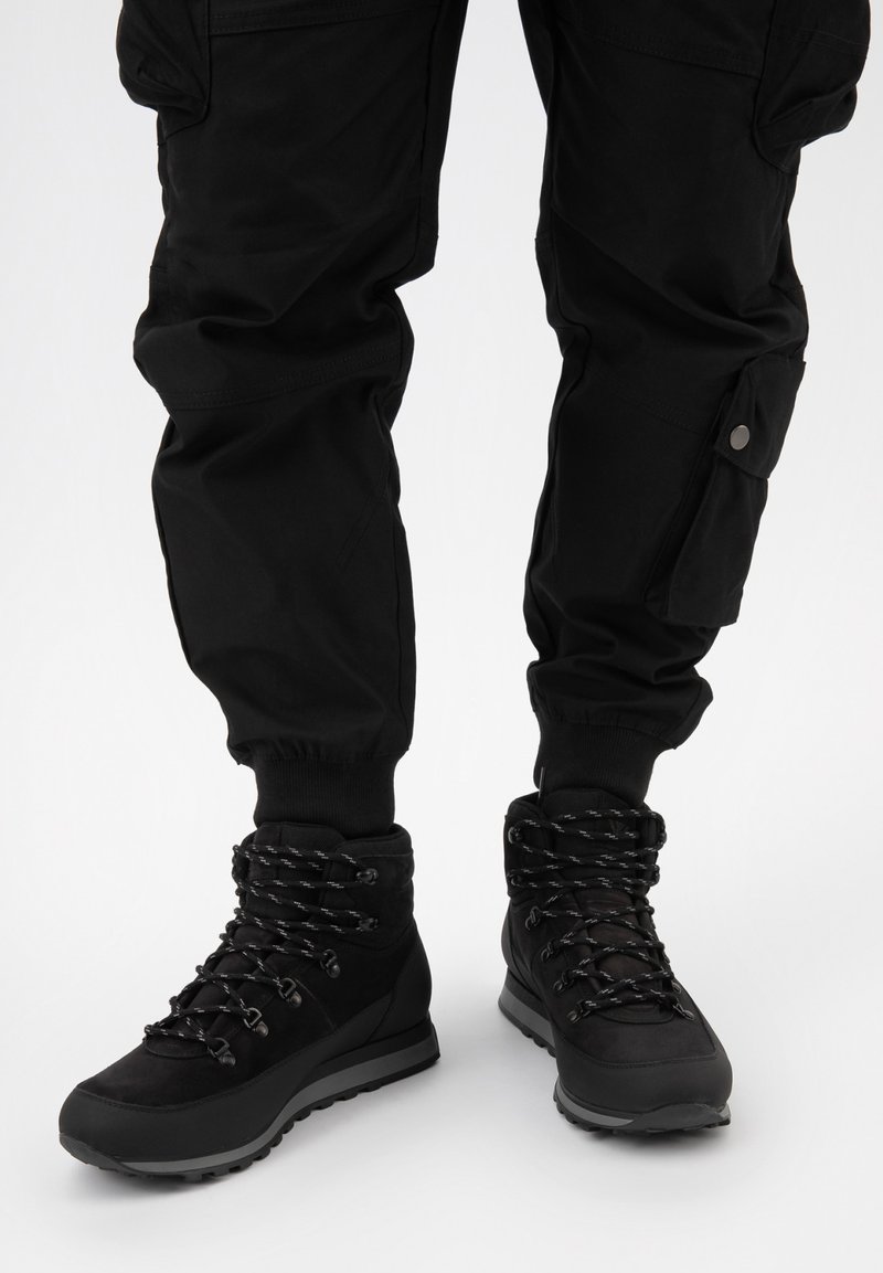 Černé turistické boty s texturovaným svrškem, zesílenou podrážkou a vzorovanými tkaničkami. V kombinaci s černými cargo kalhotami s bočními kapsami.