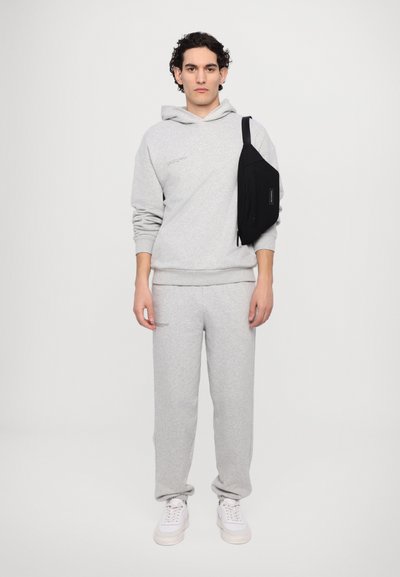 Haut en coton gris avec une poche avant et un texte subtil, associé à un pantalon de jogging gris assorti. Sac bandoulière noir sur l'épaule droite. Baskets blanches.