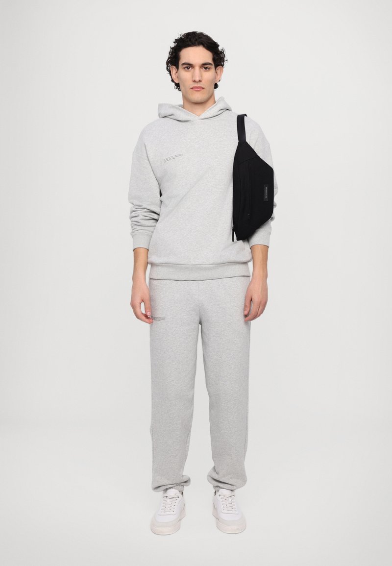 Grå bomulds hoodie med lommer foran og subtil tekst, parret med matchende grå sweatpants. Sort crossover taske over højre skulder. Hvide sneakers.