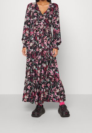 Kobieta w czarnej, kwiatowej sukience maxi z długim rękawem i wiązanym paskiem w talii, różowych skarpetkach i czarnych wsuwanych butach, stojąca na tle gładkiej ściany.