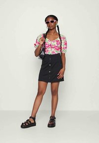 Mulher a usar uma blusa de mangas bufantes florais, saia preta com botões, sandálias pretas, óculos de sol cor-de-rosa e a segurar uma carteira preta.
