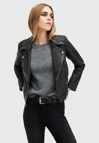 Giacca biker nera in pelle con dettagli imbottiti sulle spalle, chiusura con cerniera argentata, indossata sopra un maglione grigio in lana. Jeans neri aderenti e una cintura nera.