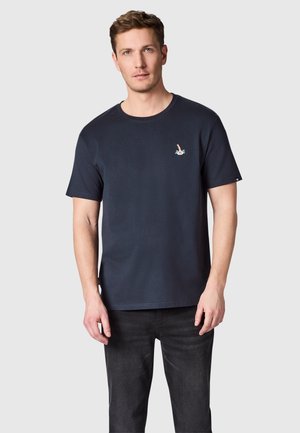Man draagt een effen marineblauw T-shirt met korte mouwen en een klein duikerslogo op de borst, gecombineerd met donkere spijkerbroek.