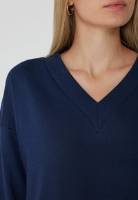 Jersey de cuello en V azul marino hecho de una tela suave. Presenta detalles acanalados en el escote, un corte relajado y un diseño minimalista.