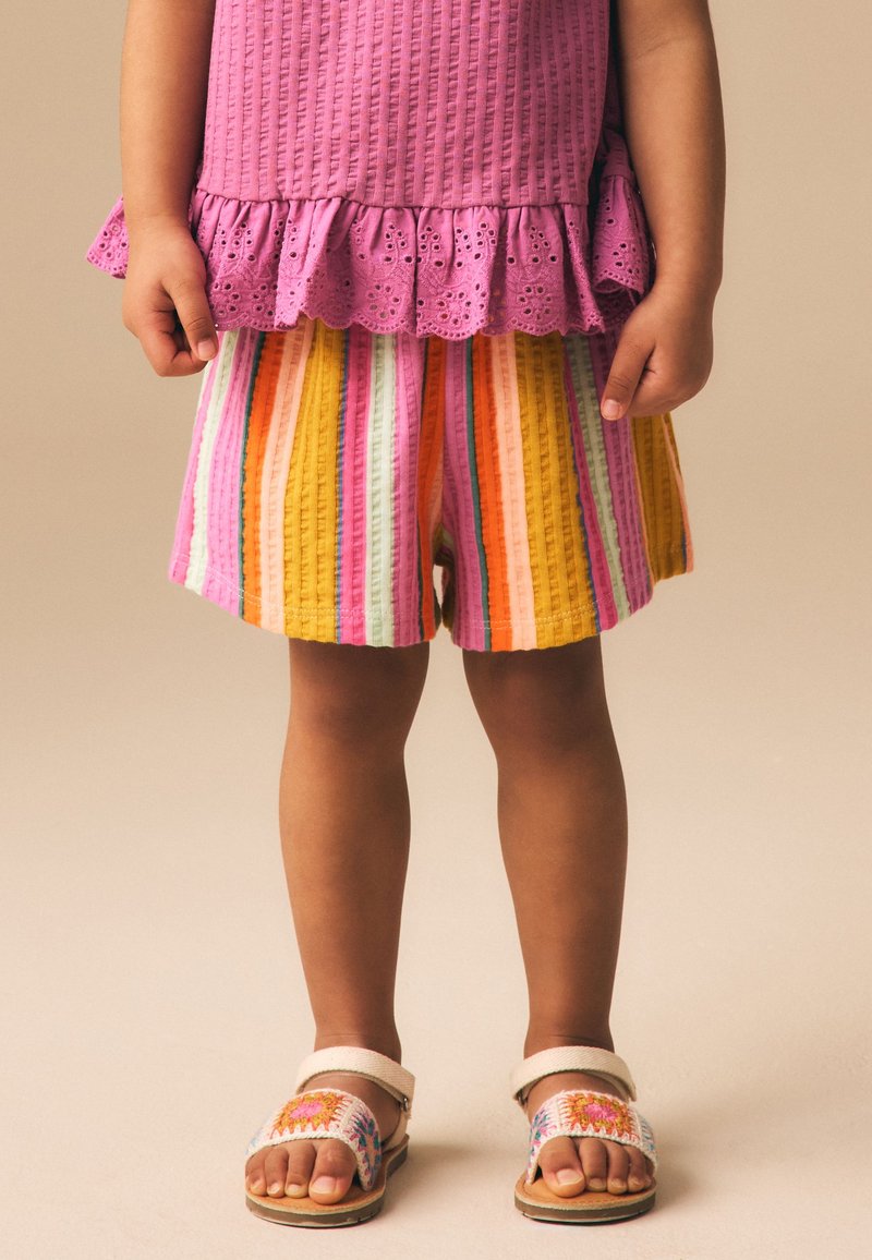 Jeune enfant portant un haut rose avec des volants, un short rayé coloré et des sandales blanches avec des motifs floraux brodés.