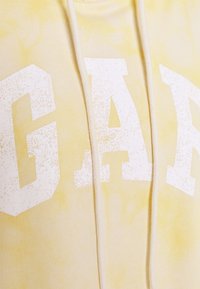 Felpa gialla tie-dye con grandi lettere bianche testurizzate "GAP" sul davanti. Presenta un cappuccio con cordini e una texture di tessuto morbido.
