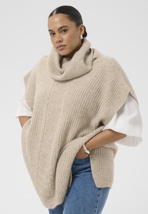 Frau trägt einen beige gestrickten Poncho mit Schalkragen über einem weißen Hemd und blauen Jeans, Hände in den Taschen, vor einem einfarbigen Hintergrund.