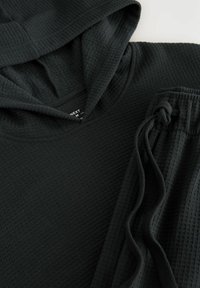 Zwarte hoodie en bijpassende broek in een gestructureerde stof met een ontspannen pasvorm. Verstelbare trekkoord in de broek. Wafelbrei ontwerp zichtbaar.