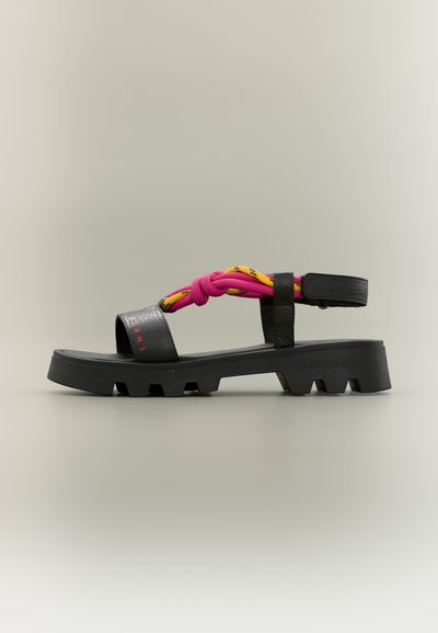 Sandale plateforme noire avec des brides nouées rose et jaune et une bride de cheville ajustable sur un fond neutre.