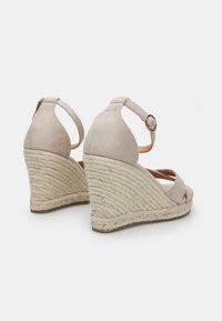 Beige suede wedge sandals with an espadrille-style rope sole, ankle strap with a metal buckle, open toe, and crisscross straps.