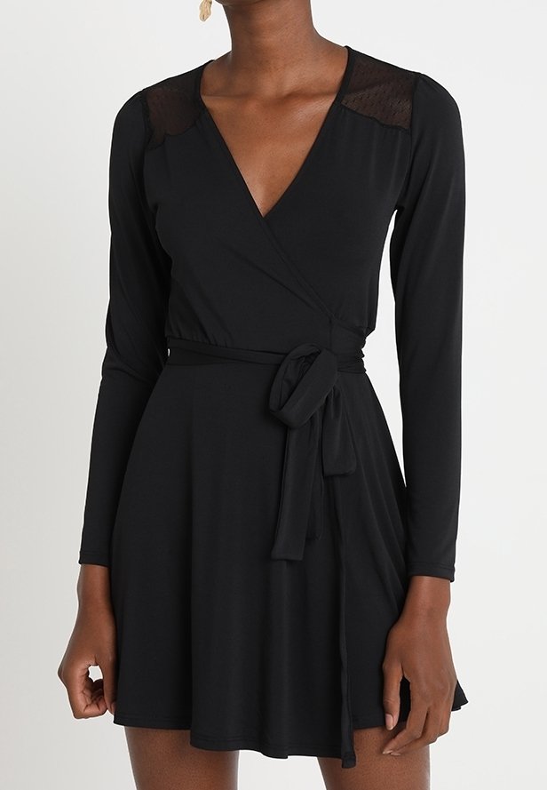 Femme portant une robe portefeuille noire à manches longues avec un détail en dentelle à l'épaule et une ceinture nouée à la taille.