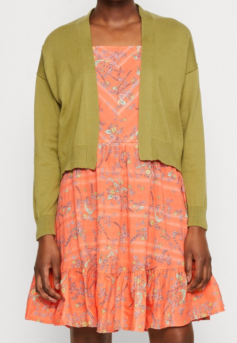 Femme portant un cardigan court vert olive sur une robe sans manches orange avec un motif floral et un ourlet à volant.