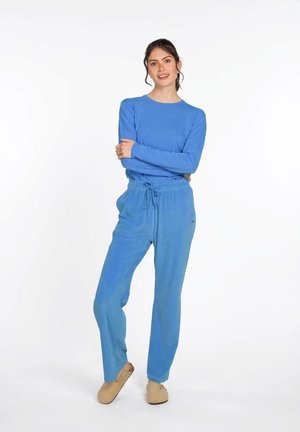 Frau steht, trägt ein blaues langärmeliges Oberteil, passende Hosen mit Kordelzug und beige Hausschuhe vor einem schlichten weißen Hintergrund.