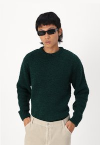 Pull en tricot texturé vert avec un col rond et des manches longues, associé à un pantalon clair et des lunettes de soleil sombres.